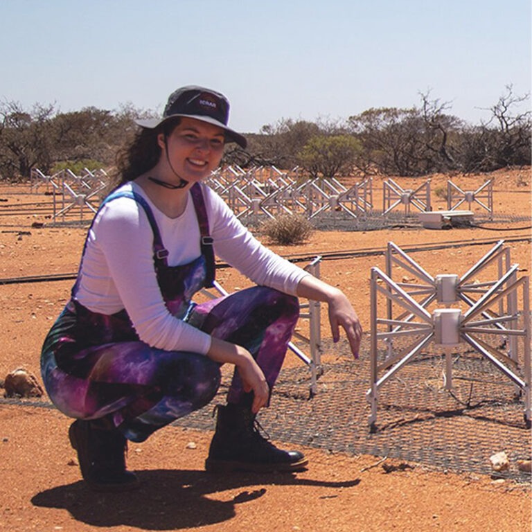 Dr Kat Ross crouches smiling next to some of the many antennas that make up the MWA telescope