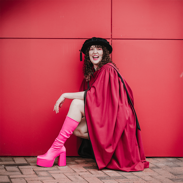 Dr Kat Ross earned her PhD in Astronomy and Astrophysics from Curtin University. She is pictured in a red graduation robe, pink heeled boots, and a black cap. She poses happily against a red wall.