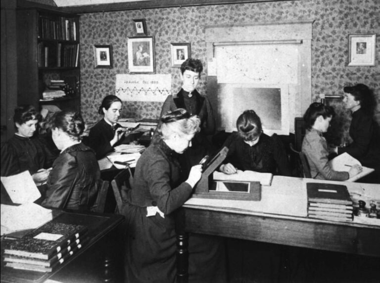 A black-and-white photograph of a group of women computers (people who performed calculations and computations) at Harvard College Observatory. The scene captures the historical essence with Victorian-style clothing, vintage technology, and wallpapered walls adorned with posters depicting mathematical equations. A group sits around wooden desks, surrounded by books.