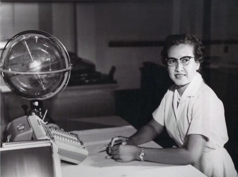 A monochrome photograph of NASA research mathematician Katherine Johnson. She wears a white dress and black-rimmed glasses and is smiling at the camera while sits at a desk.