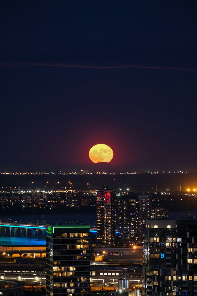 A large yellow Moon rises over a city at night