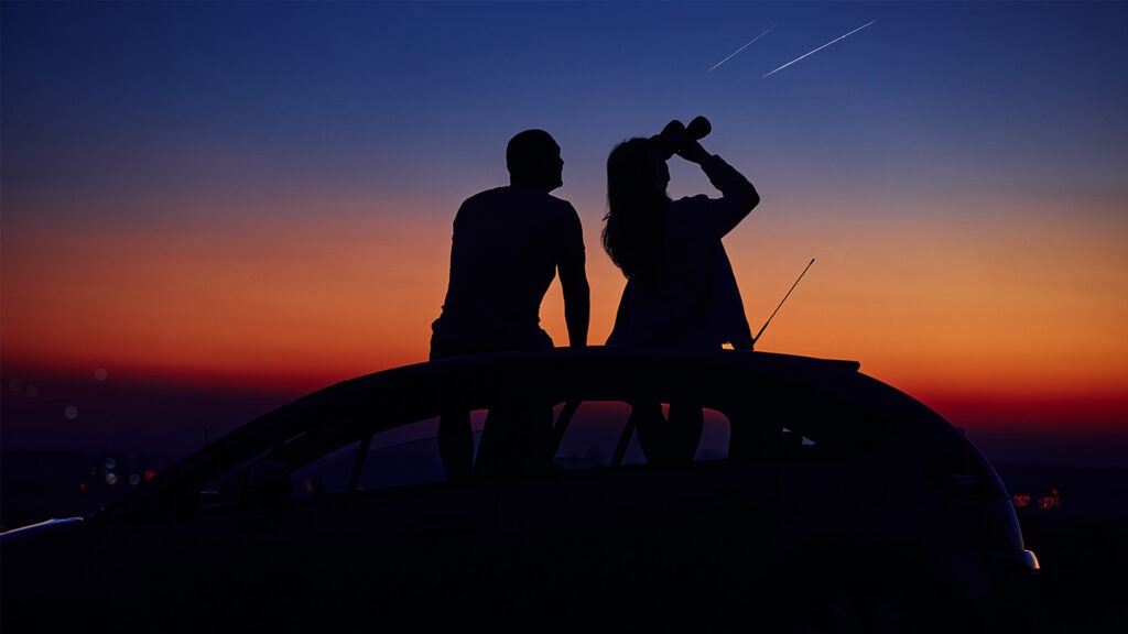 A couple sit on the roof of a car watching a meteor shower through binoculars.
